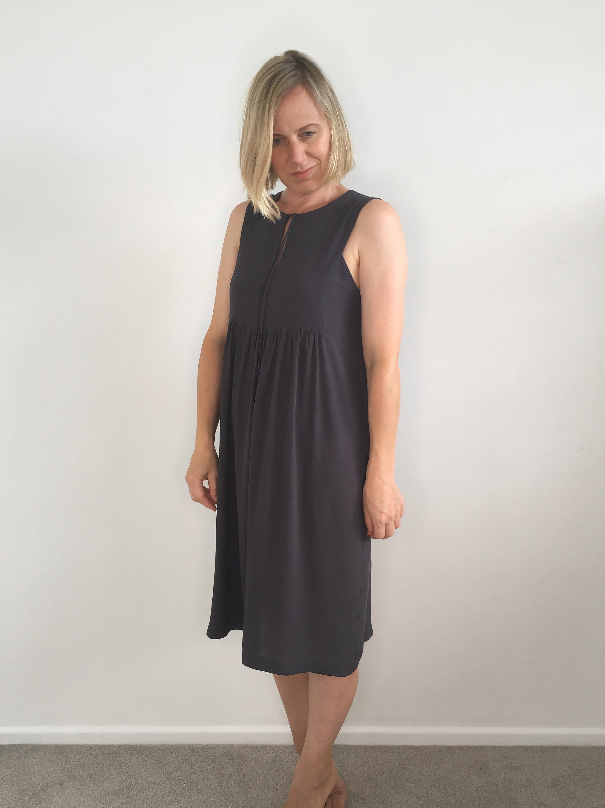 A woman wears a knee-length dark grey sleeveless viscose dress with front tie & gathered skirt.