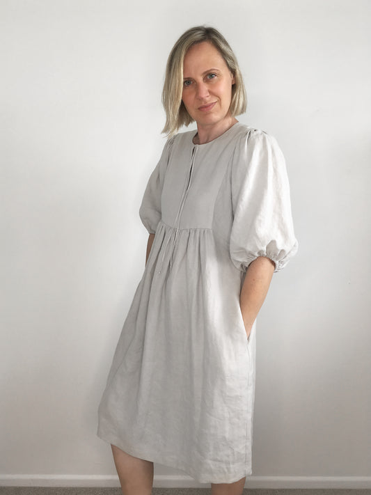 A woman wears a knee-length  pale grey linen dress with voluminous sleeves and gathered skirt