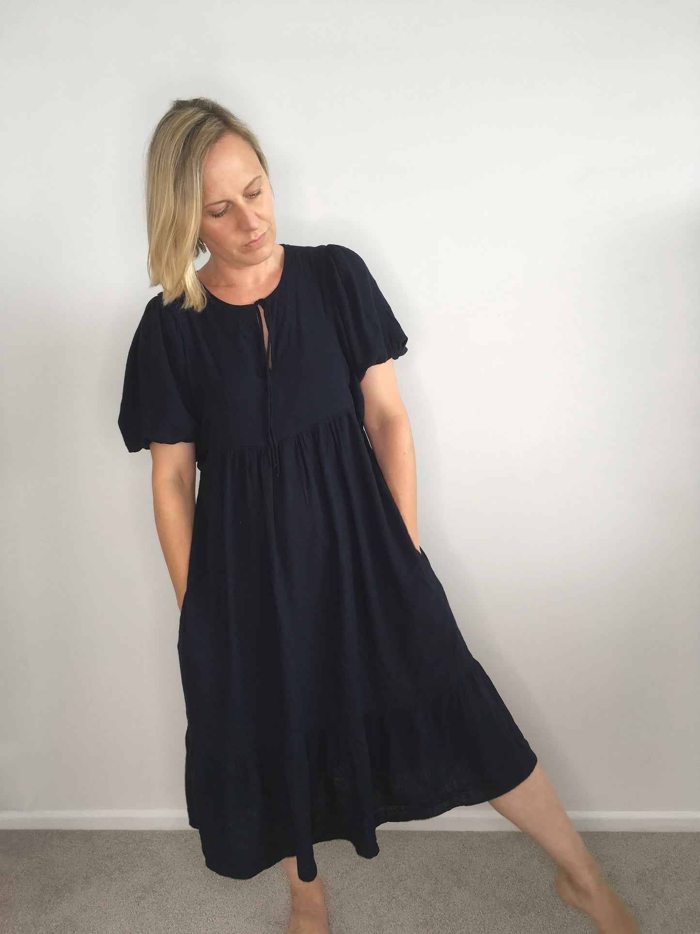 A woman wears a navy viscose dress with short puff sleeves, front tie at the neck & gathered skirt. A ruffle hem panel finishes below the knee