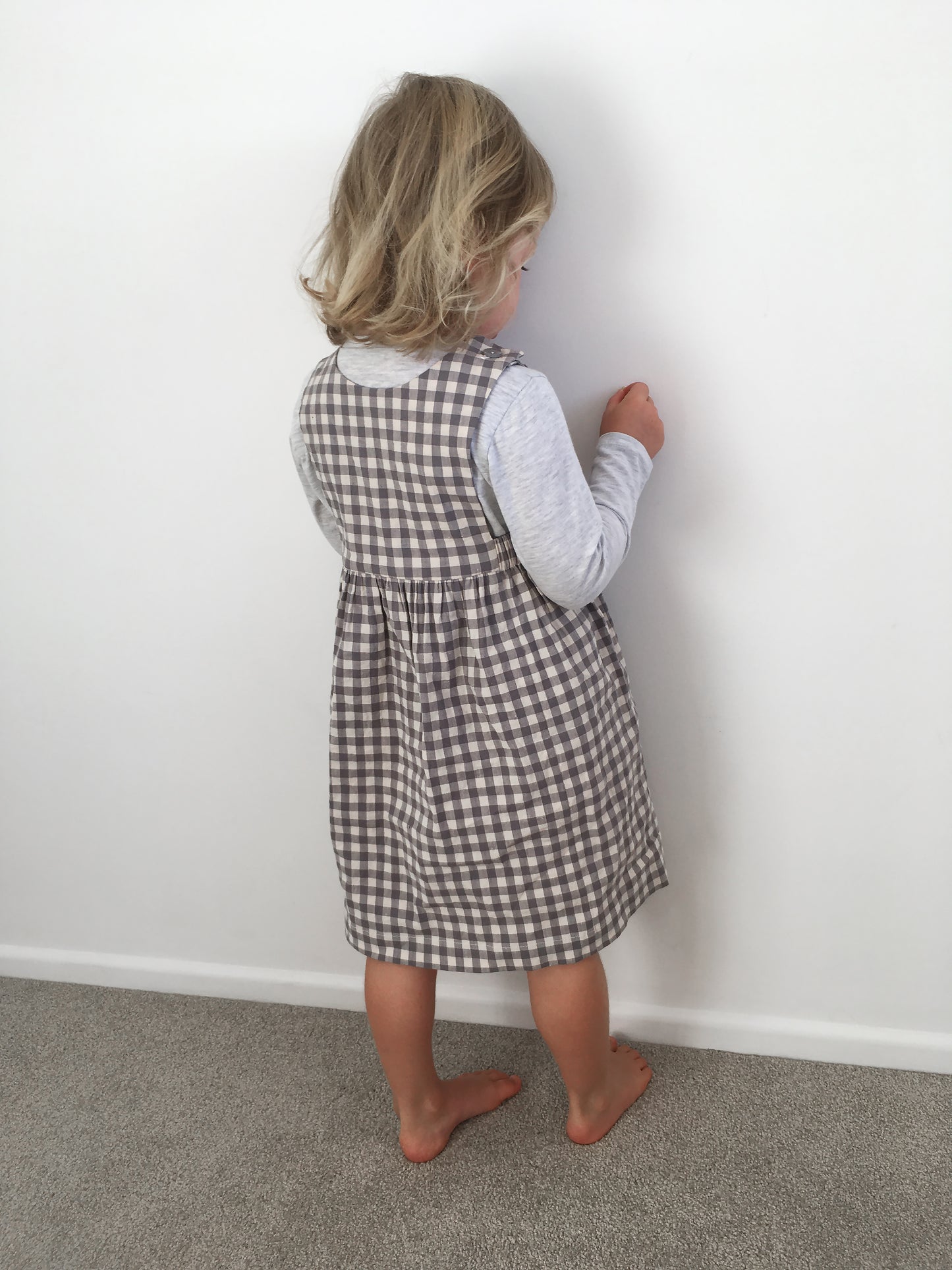 A small girl scratching a pristine white wall with a scrunched up easter egg wrapper. Wears a gingham linen pinafore over a light grey long sleeved tee.