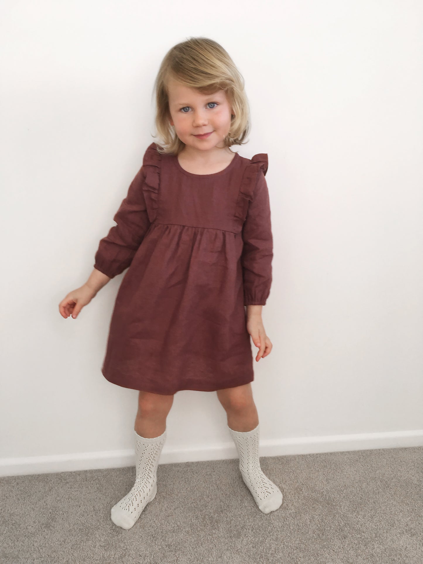 A small girl wearing a burgundy coloured linen dress with ruffle detail on the bodice